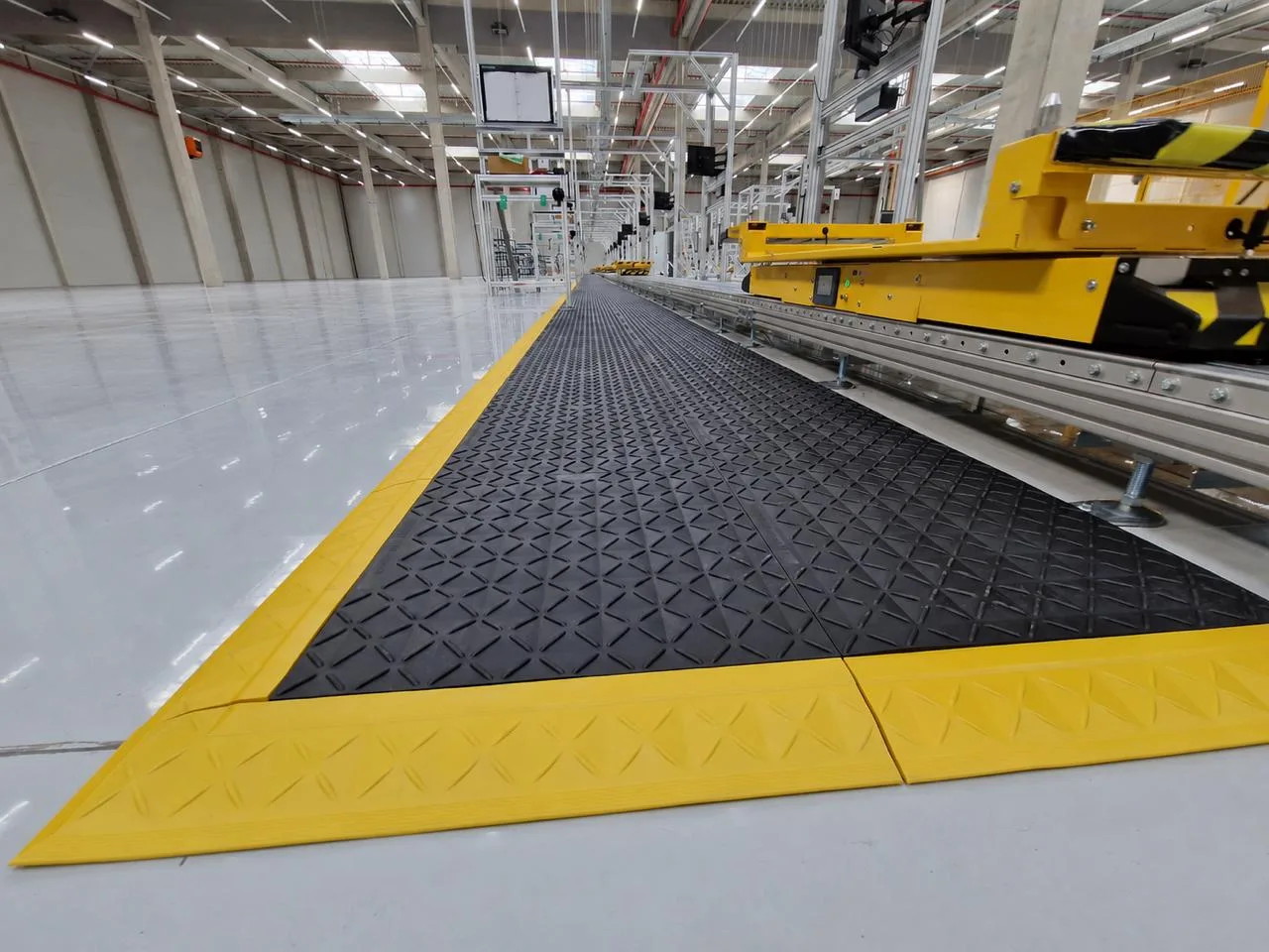 Black modular anti-fatigue matting with yellow safety edging running alongside an automated conveyor line in a warehouse