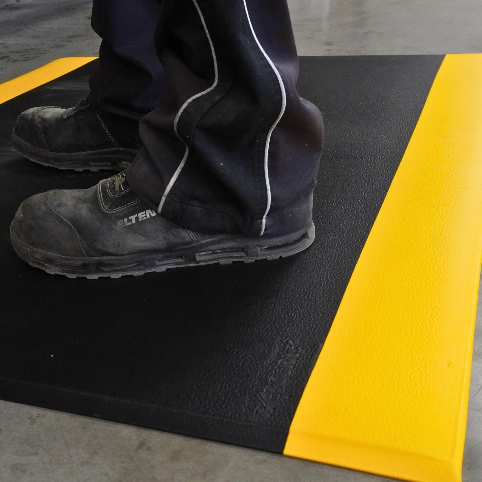 Close-up of safety boots on an M1 black and yellow anti-fatigue mat on a concrete floor